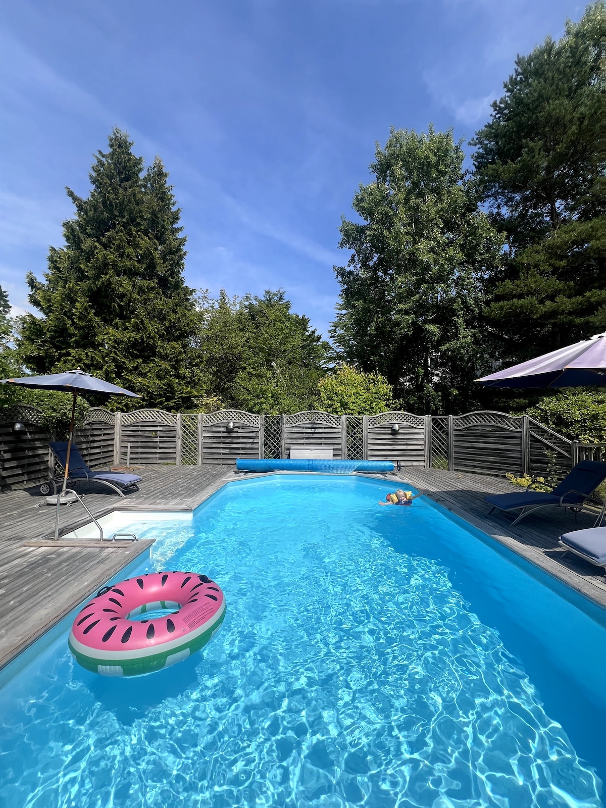 A serene backyard pool area is surrounded by tall trees and a wooden deck. Two umbrellas provide shade for lounge chairs positioned around the pool. A pink inflatable float adds a pop of color to the clear blue water, inviting relaxation.