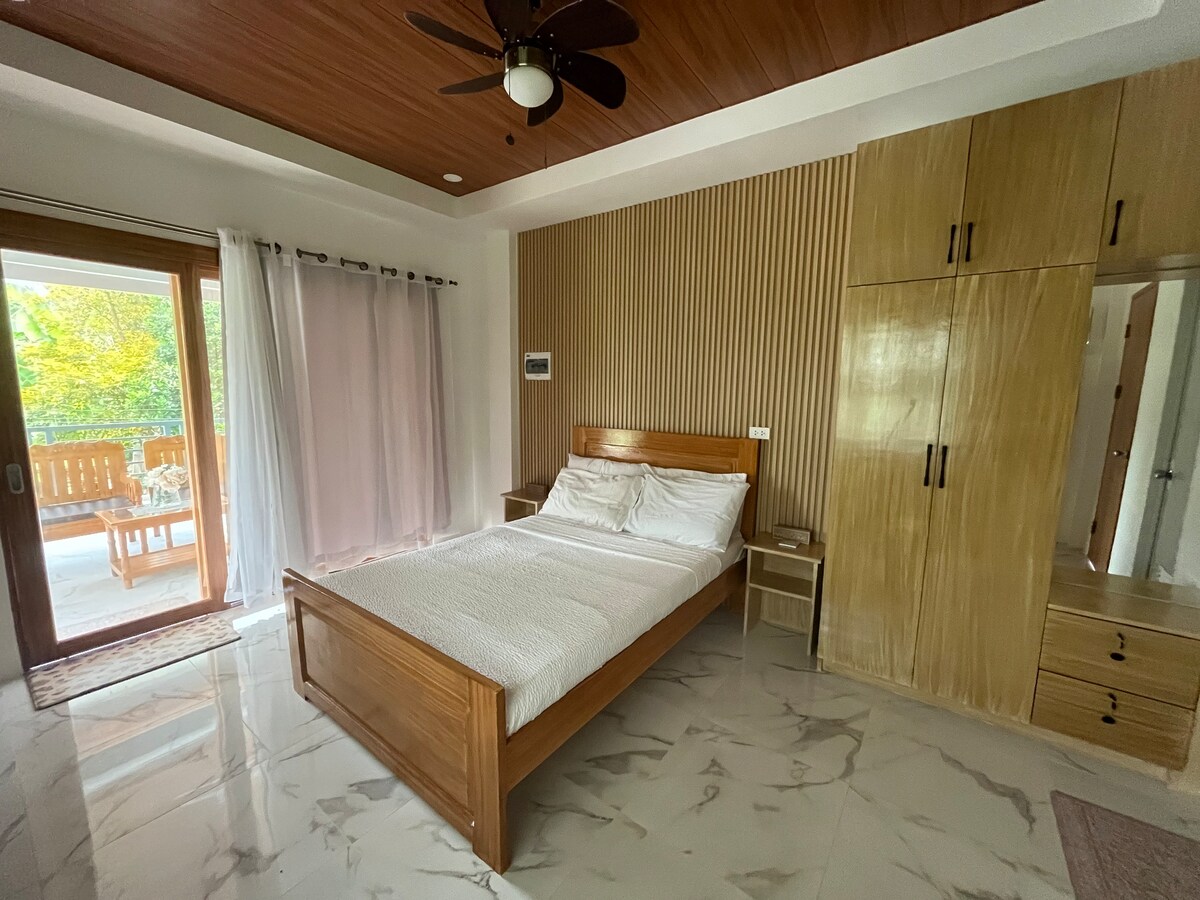 A bright bedroom features a wooden bed with a soft white bedding set. Light marble flooring complements the space. A tall wardrobe and bedside table are present, while large glass doors lead to a furnished outdoor area, allowing natural light to fill the room.