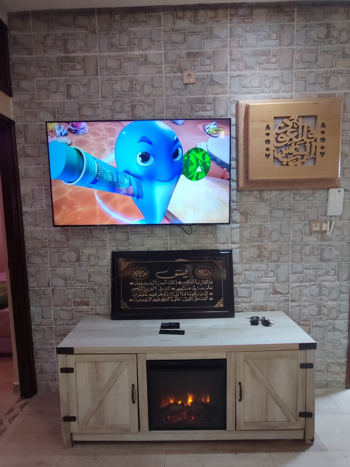 A flat-screen television is mounted on a textured wall, displaying a colorful animated character. Below, a rustic fireplace adds a cozy touch to a light-colored media cabinet, which contains decorative items and an inscription on a framed piece above it.