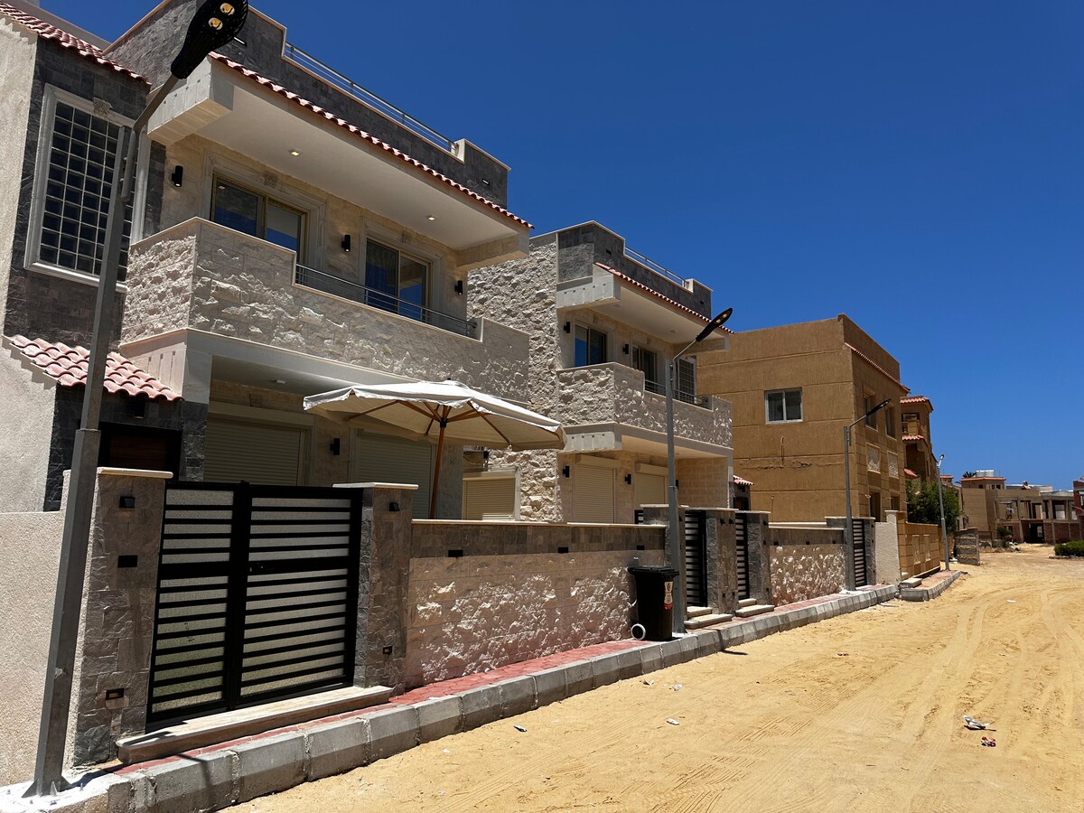 Two modern homes are depicted side by side, featuring stone facades and red-tiled roofs. An umbrella stands open in front of one home, while a sandy path leads between the properties, framed by clear blue skies. Streetlights and a fence are visible, adding to the structure's appeal.