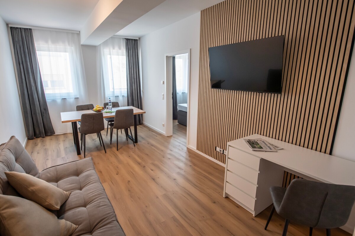 A modern living area features a comfortable sofa facing a wall-mounted television. A dining table surrounded by four chairs is positioned nearby, while a work desk with a chair is placed against the wall. Natural light filters through two windows, enhancing the warm wooden flooring.