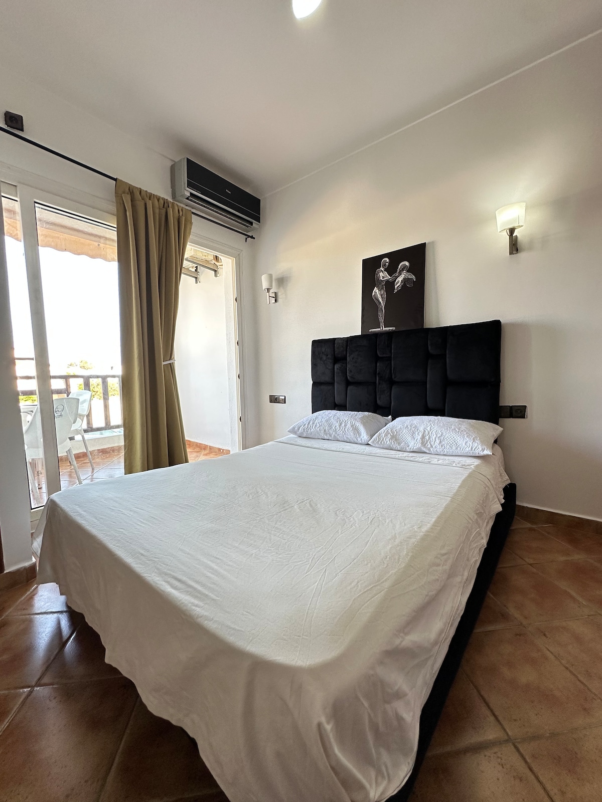 A spacious sleeping area features a large bed with a minimalist headboard. Natural light enters through sliding glass doors that lead to a balcony. Soft curtains frame the entrance, and wall sconces provide additional lighting. A piece of wall art is displayed above the bed.