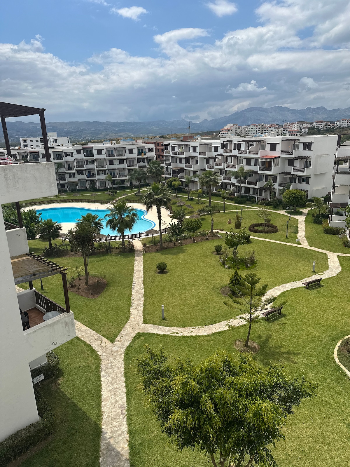 A well-maintained garden area is presented, featuring pathways that meander through manicured lawns and clusters of trees. A swimming pool is visible in the center, surrounded by tropical landscaping. Several benches are scattered throughout the space, providing serene spots to relax.