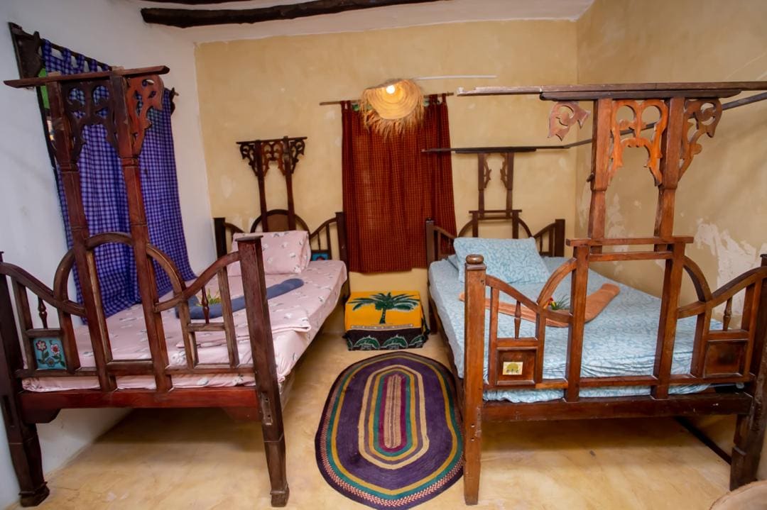 A cozy sleeping area is displayed with two wooden beds, featuring intricate headboards. Textiles in soft colors adorn the beds, complemented by a patterned area rug. Natural light enters through the window, covered by a checkered curtain, enhancing the room's inviting design.