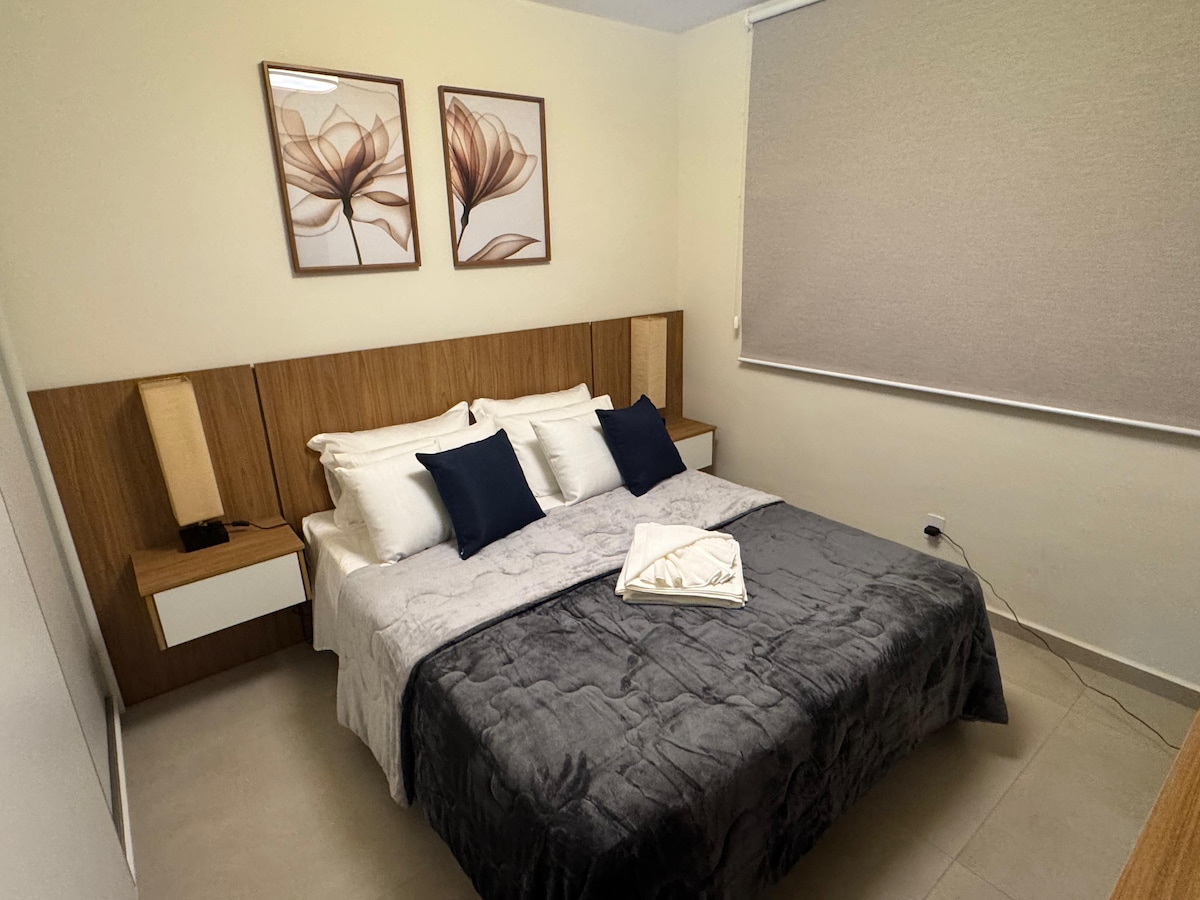 A comfortable sleeping area features a spacious bed with soft linens and pillows. Simple wooden furnishings, including a headboard and nightstands with lamps, complement the serene decor. Two floral art pieces adorn the wall, adding a subtle touch to the ambiance.