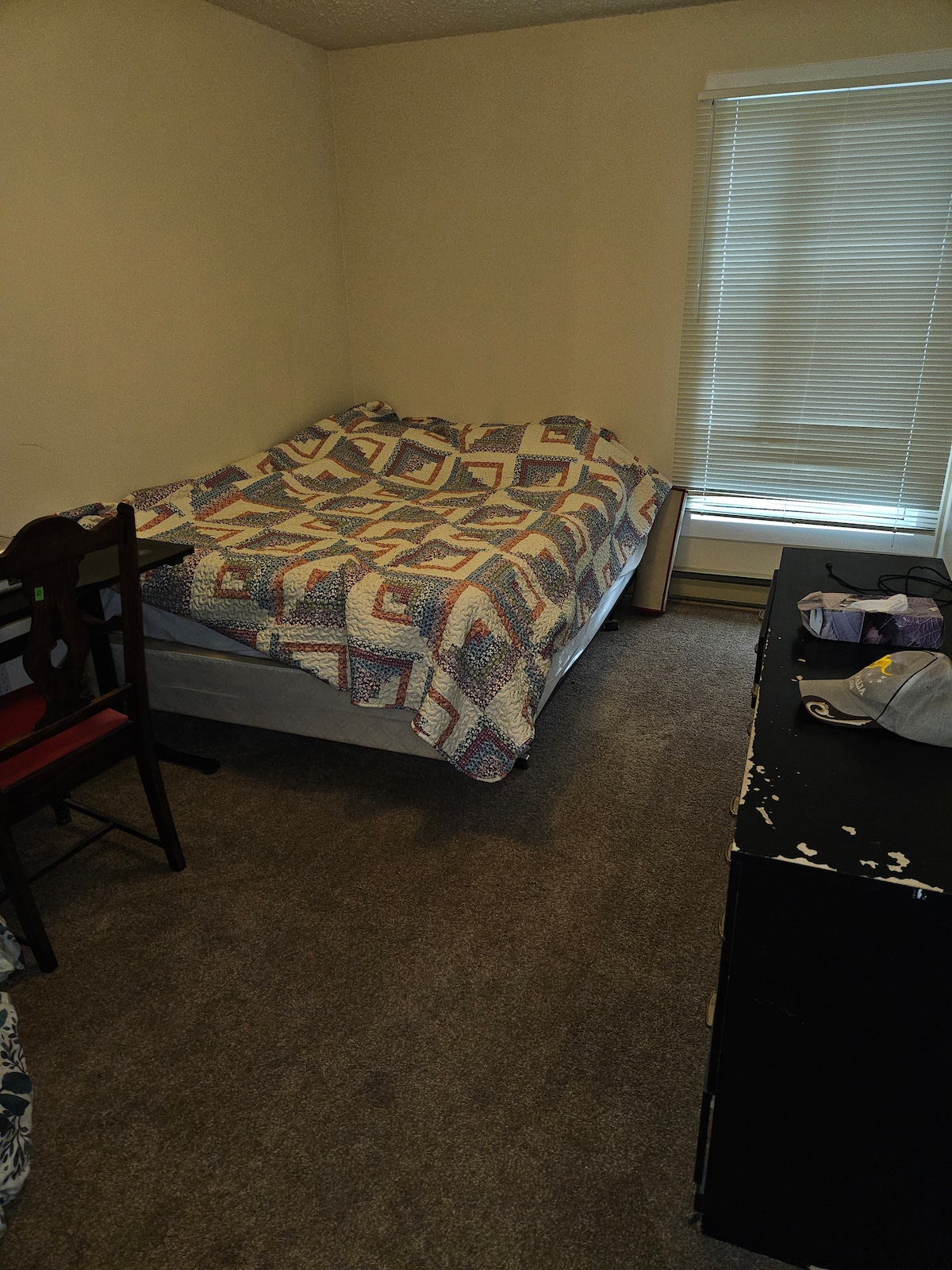 A compact bedroom is furnished with a spacious bed draped with a patterned quilt. A chair is positioned nearby, and a dresser is visible in the corner. Natural light filters through a window covered by blinds, casting soft shadows across the carpeted floor.