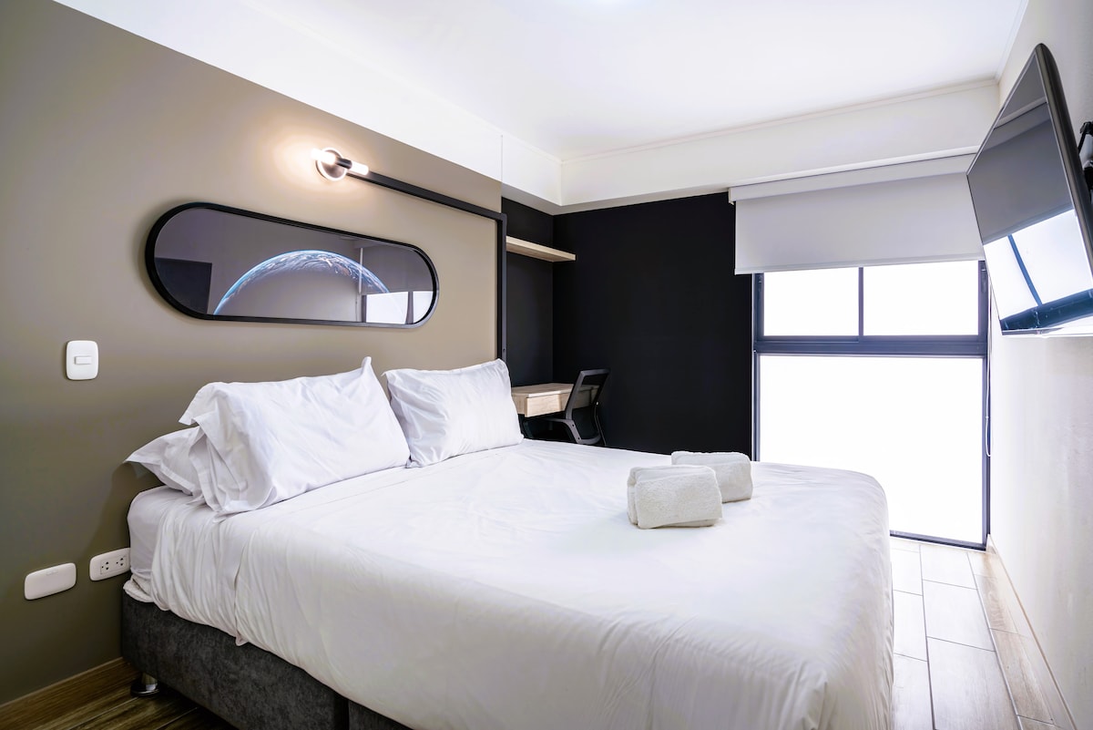 A spacious bedroom features a large bed with fresh white bedding and two rolled towels. A sleek desk with a chair is visible, alongside a wall-mounted television. Natural light filters in through a window, which is partially covered by a blind, with a dark accent wall adding depth.