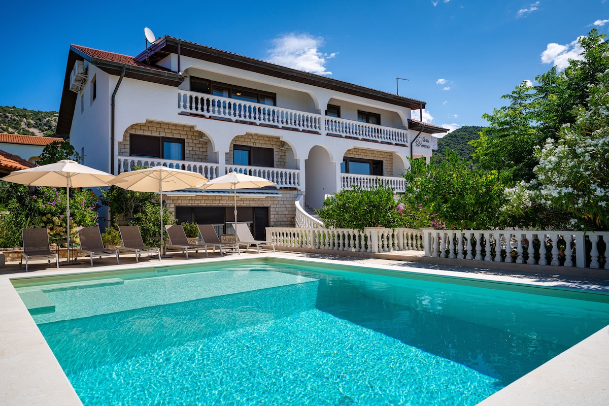 The exterior of the villa is showcased with a heated outdoor pool in the foreground, reflecting the clear sky. Sun loungers are arranged neatly along the poolside, while a spacious terrace features umbrellas and green foliage adds to the serene setting.