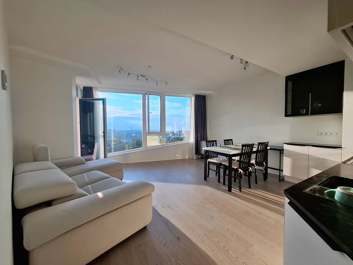 A spacious living area features large windows that provide a panoramic view of the Tallinn skyline and gulf. A light-colored sectional sofa sits in one corner, while a dining table with seating for six is positioned centrally. Smooth wooden flooring enhances the bright environment.