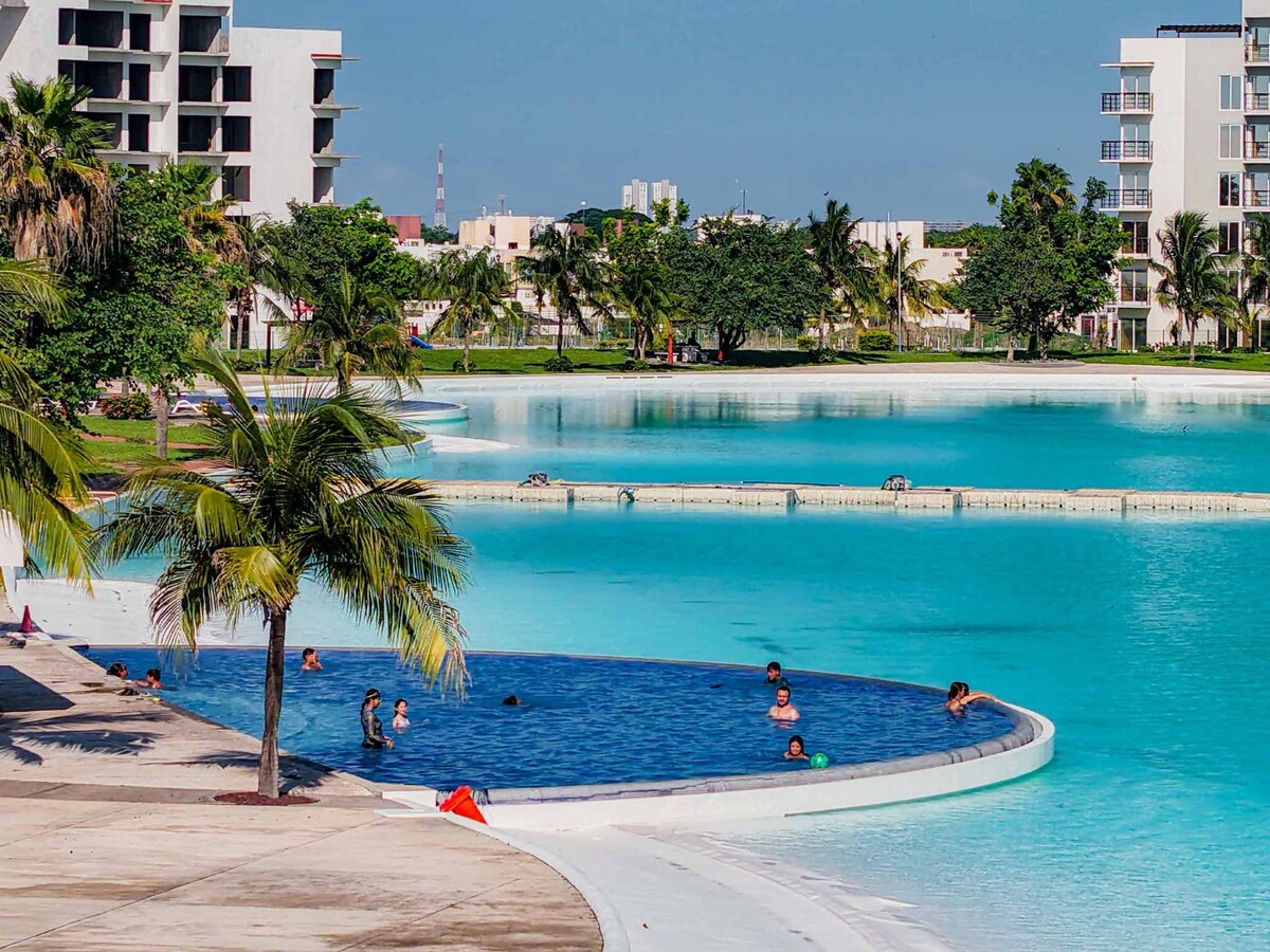 A large artificial lagoon is featured, surrounded by lush green areas and palm trees. Calm turquoise waters lap at the edge, where families and individuals can be seen enjoying recreational activities. Distinctive modern buildings are visible in the background, enhancing the serene environment.