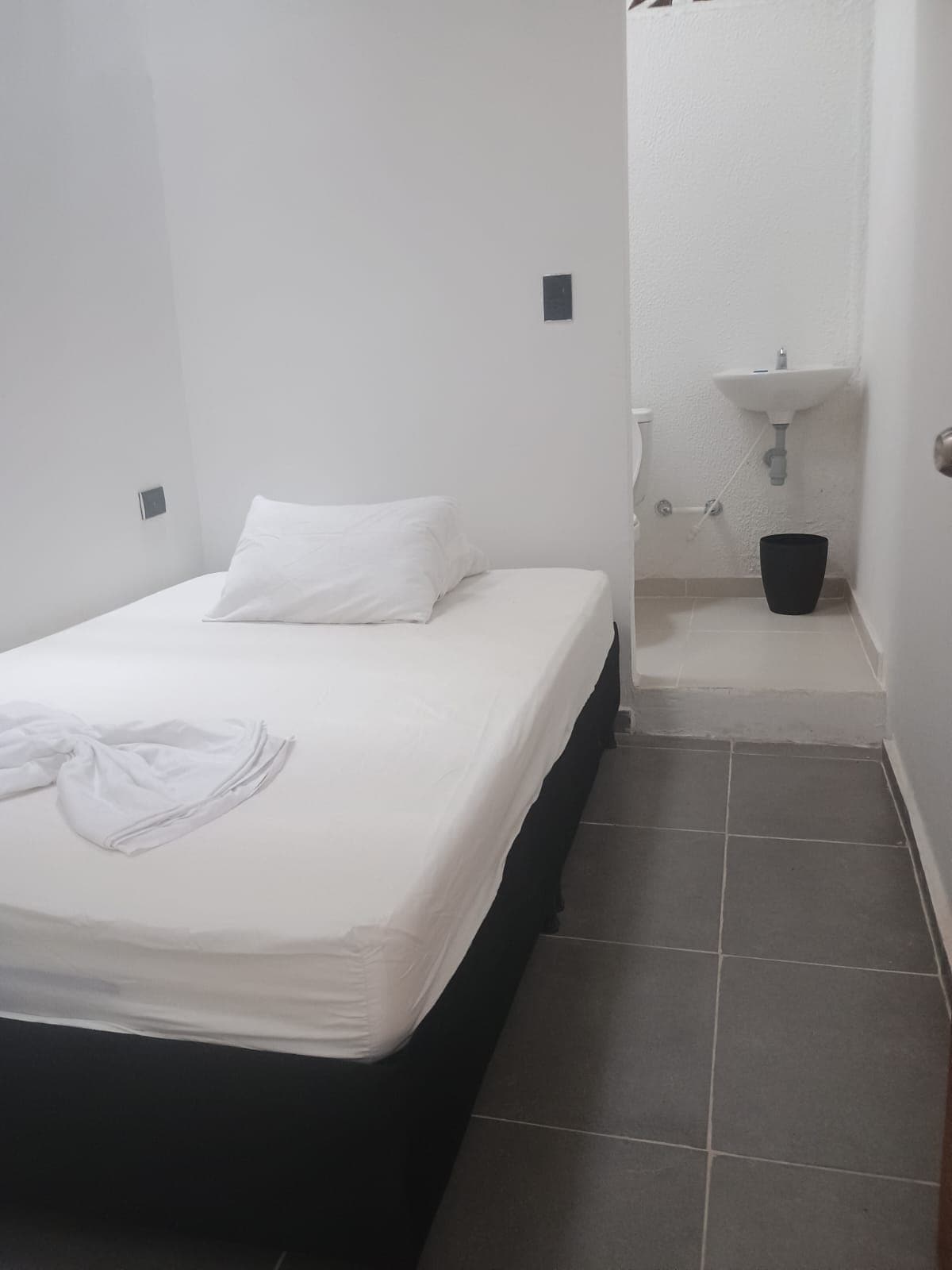 A modest sleeping area features a spacious bed with plain white linens and a decorative towel. The floor is covered with gray tiles, and a small bathroom area is visible in the background, showcasing a sink and toilet within a minimalistic setting.