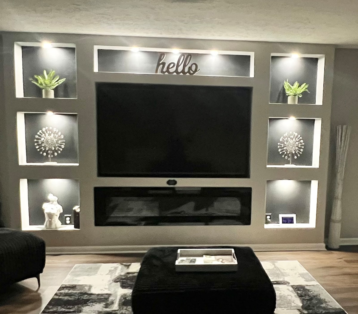 A modern entertainment area features a large wall-mounted television situated above a sleek electric fireplace. Decorative shelves are illuminated, displaying plants and geometric accent pieces. A dark, textured rug anchors the space, while a light-colored tray is positioned on a low table in front.