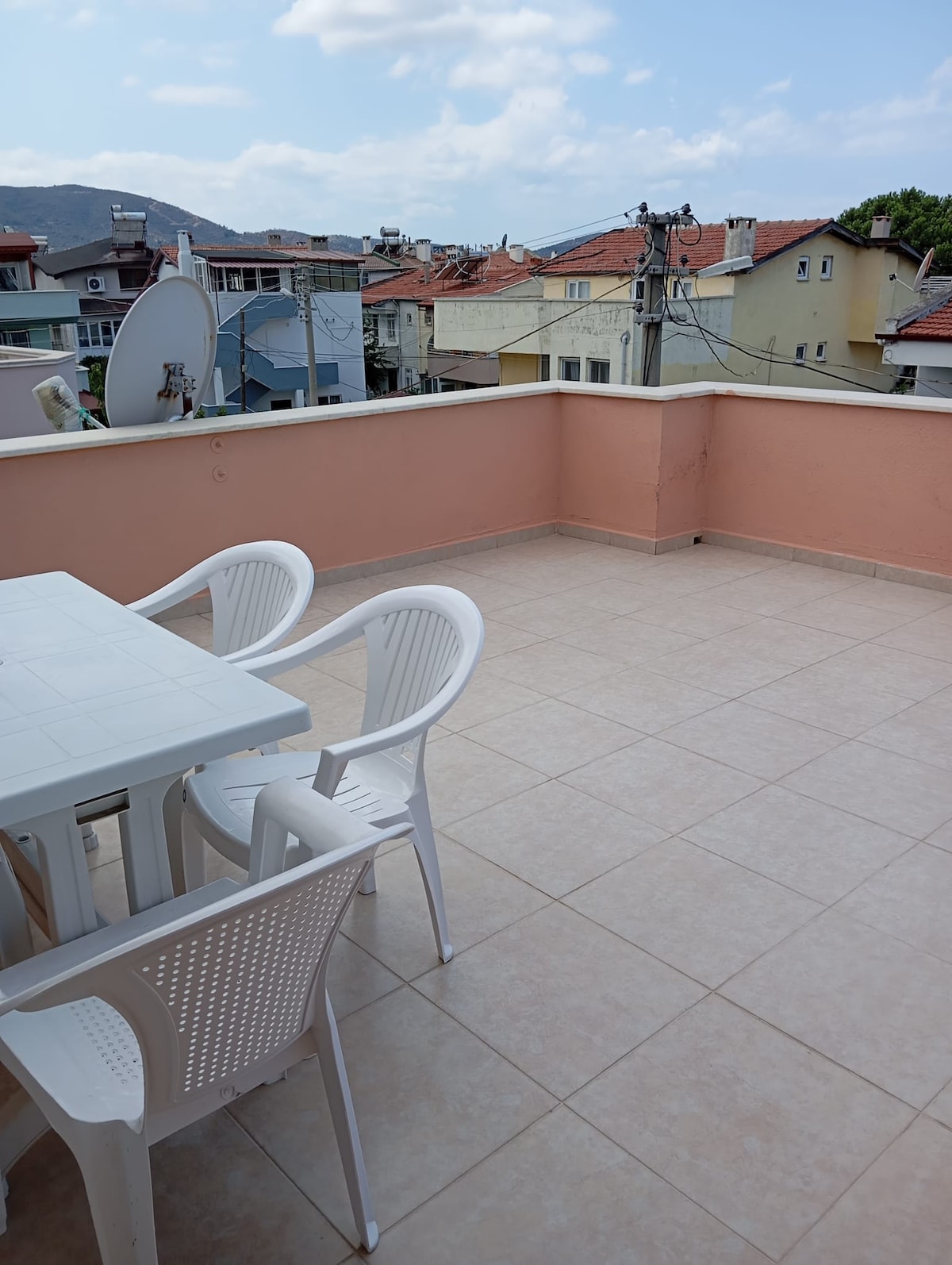 A spacious terrace features a white table and two white chairs, set against a backdrop of rooftops and distant mountains. The tiled floor provides ample room for relaxation. A satellite dish is visible on a nearby building, and the area is bathed in natural light.