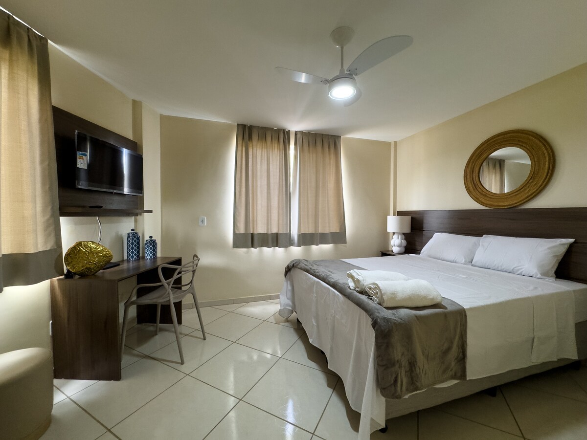 A well-appointed suite features a spacious bed with crisp linens and soft pillows. Natural light filters through the large windows, complemented by neutral-toned curtains. A small desk and chair are positioned next to the bed, while a round decorative piece and a mirror add subtle elegance.