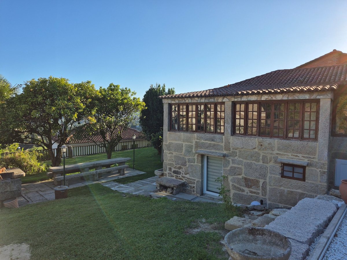 A charming stone house features large windows that allow natural light to fill the interior. A spacious patio area is shown, complete with a long table and benches, surrounded by green grass and trees. The landscape indicates a peaceful outdoor environment.