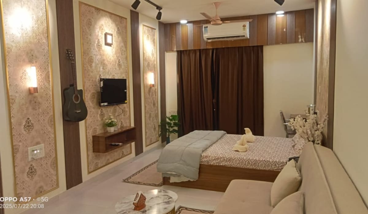 A cozy studio apartment features a spacious bed with soft linens and decorative throw pillows. A comfortable sofa is positioned next to a marble coffee table. Wall-mounted decor includes a guitar and a small television, with ambient lighting enhancing the serene atmosphere.