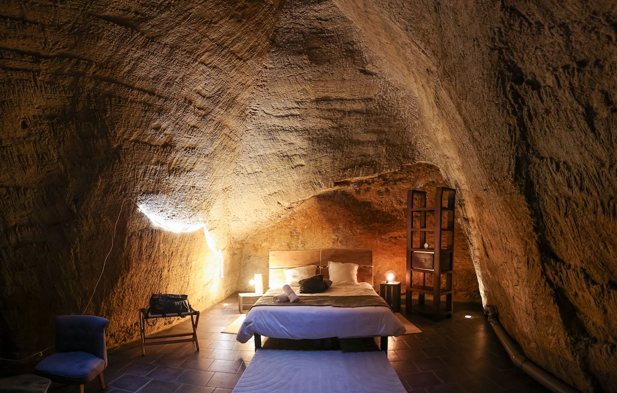 A spacious sleeping area is set within a troglodyte structure, featuring a large bed against a textured stone wall. Soft lighting from lamps creates a warm ambiance. A small chair is positioned nearby, adding to the cozy and inviting environment.
