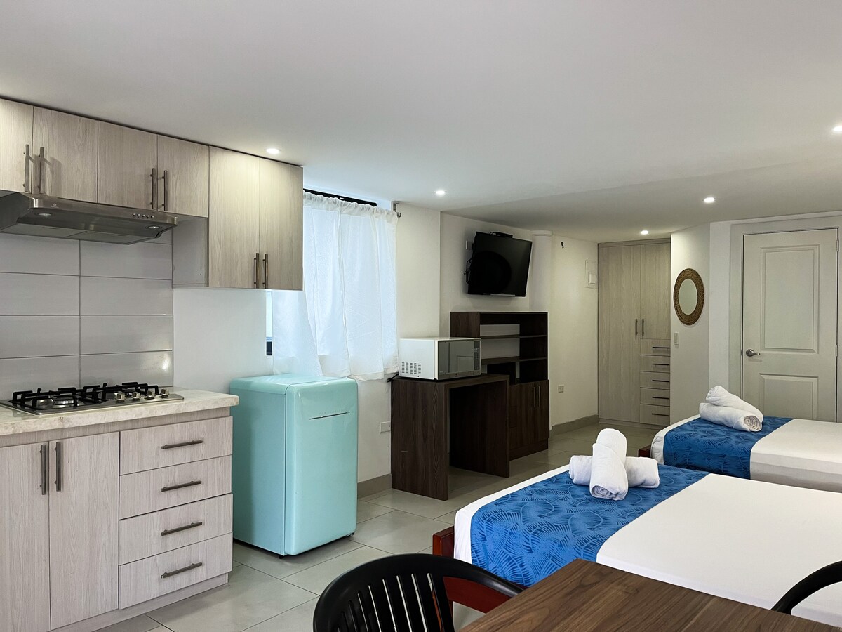 A contemporary suite features a compact kitchen area with light wood cabinetry and a retro-style refrigerator. Two beds are dressed in blue linens, and a dining table is set nearby. A flat-screen television is mounted on the wall, enhancing the functional layout of the space.