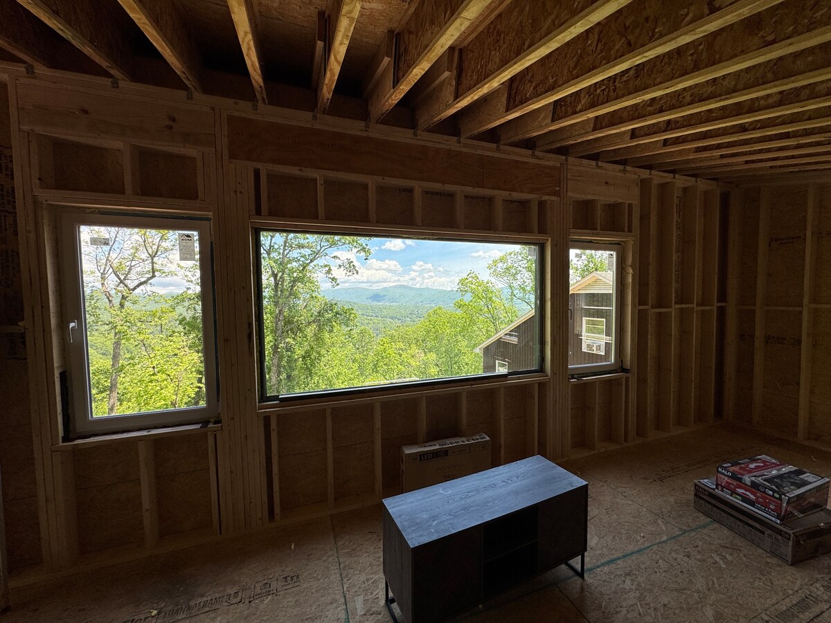 A large picture window frames an expansive mountain view, allowing natural light to fill the space. The unfinished walls and ceiling showcase the raw structure, while a simple coffee table rests in the center of the room. The openness evokes a sense of spaciousness.