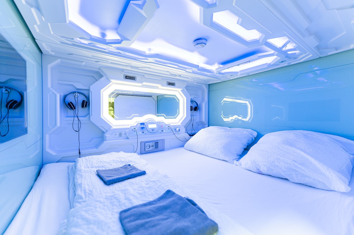 A modern sleeping pod features sleek blue walls and ambient lighting. Soft white bedding creates a clean and serene environment. Two pillows are neatly arranged on the spacious bed, with towels placed nearby. Windows allow natural light to fill the pod, enhancing the overall atmosphere.