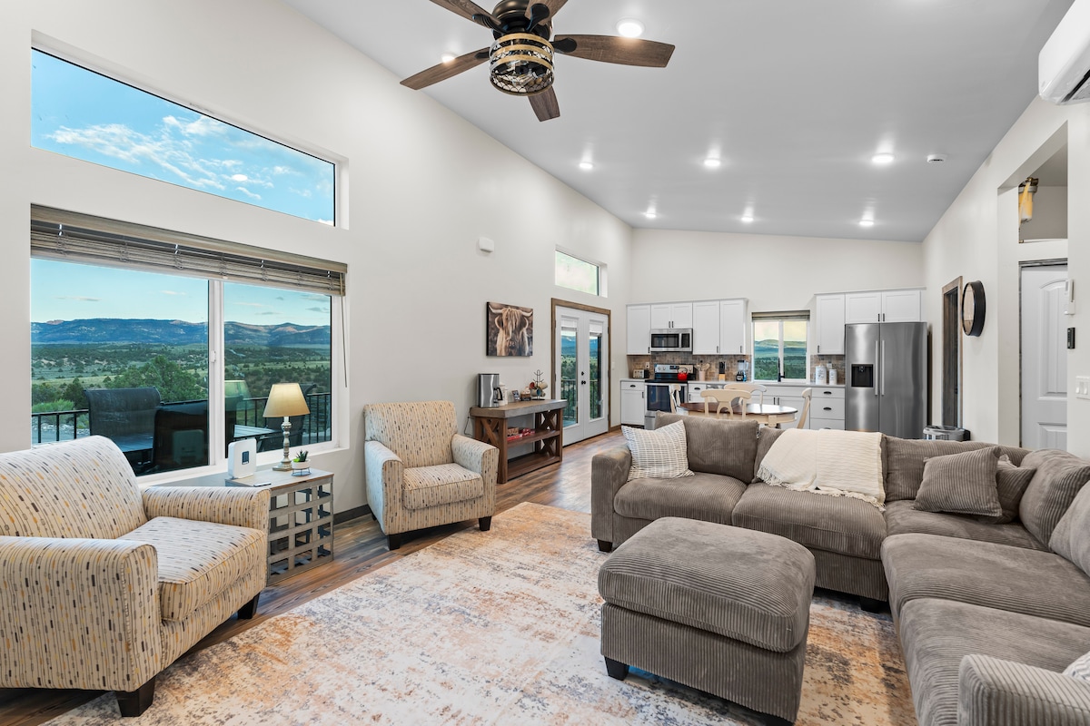 A well-lit open living space features a large sectional sofa and two armchairs placed near a window, offering expansive views of the surrounding mountains. A coffee table rests on a light area rug, while a kitchen area with modern appliances is visible in the background.