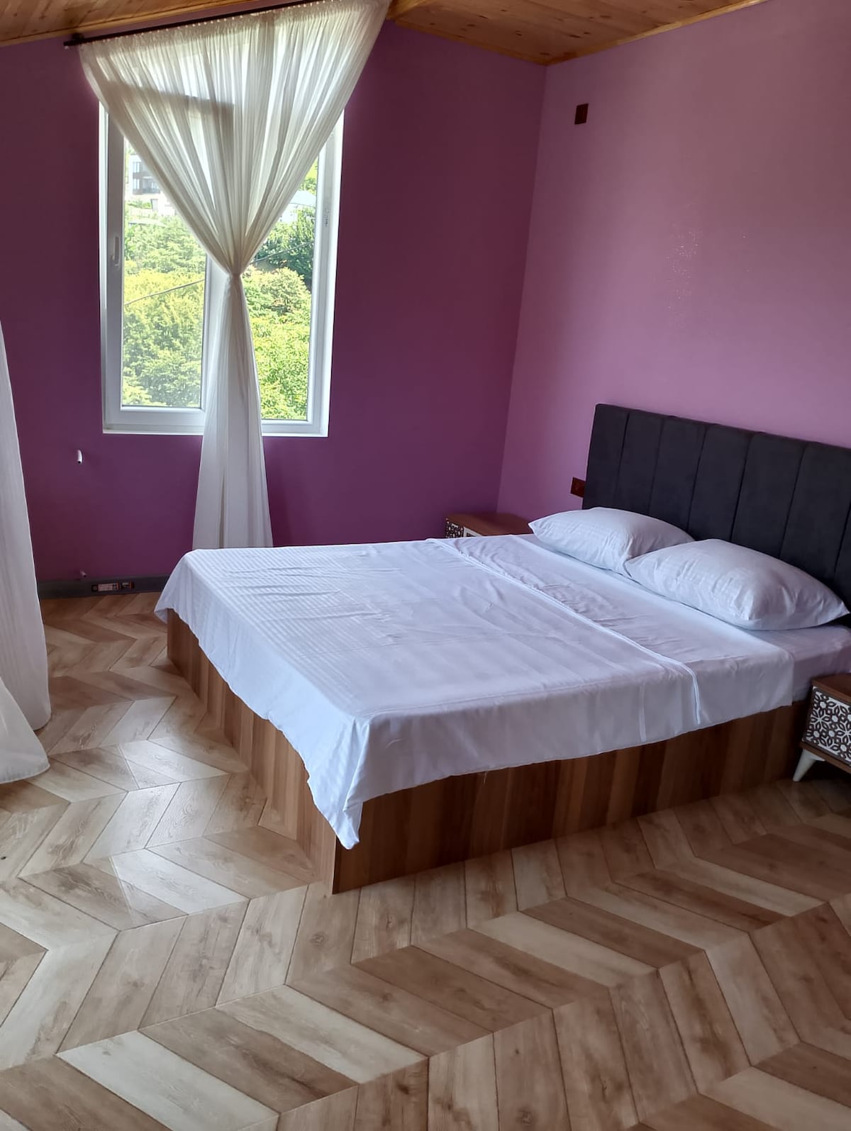 A sleeping area features a spacious bed with crisp white linens and fluffy pillows. The room's light pink walls create a bright atmosphere, and a large window offers views of the surrounding greenery. Light wooden flooring enhances the space's natural feel.