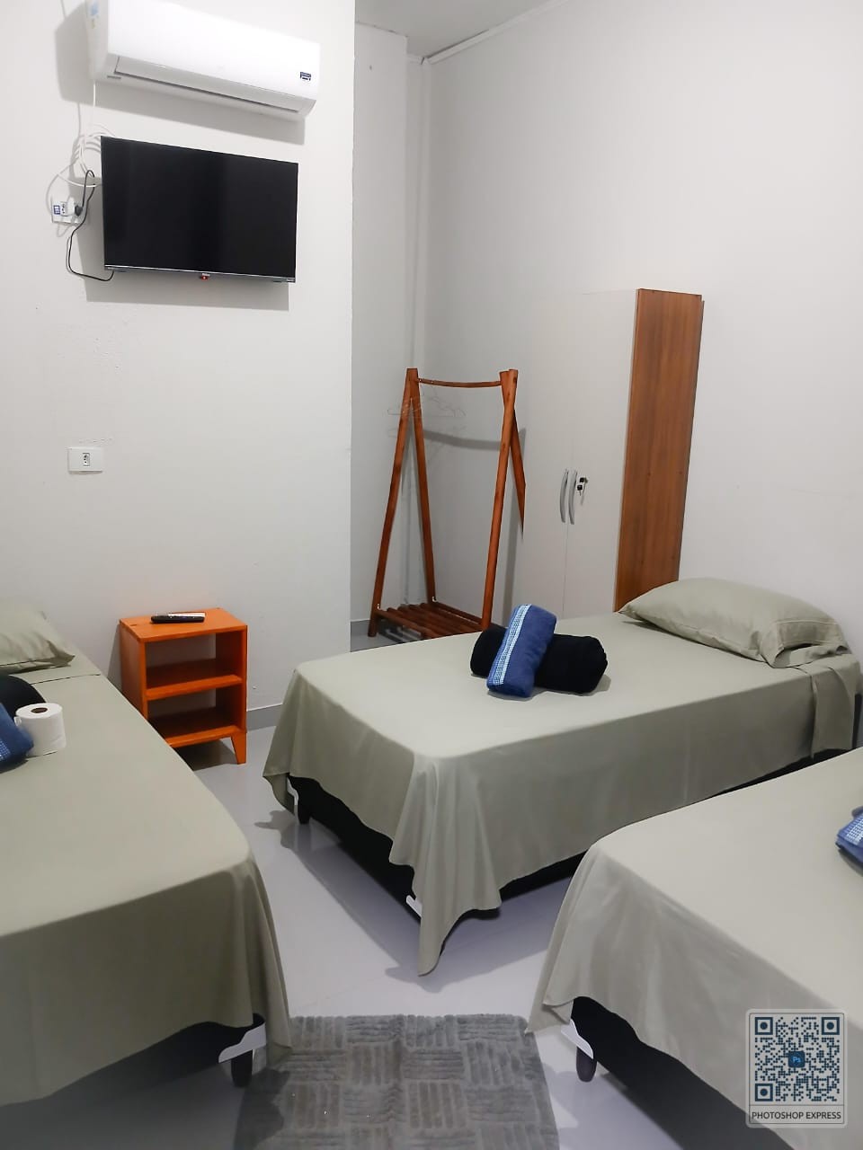 A well-organized room features three twin beds dressed in light grey bedding. A small orange shelf is positioned beside one bed, while a wooden clothes rack and a flat-screen TV are mounted on the wall. A neutral-colored rug is placed on the floor, adding comfort.