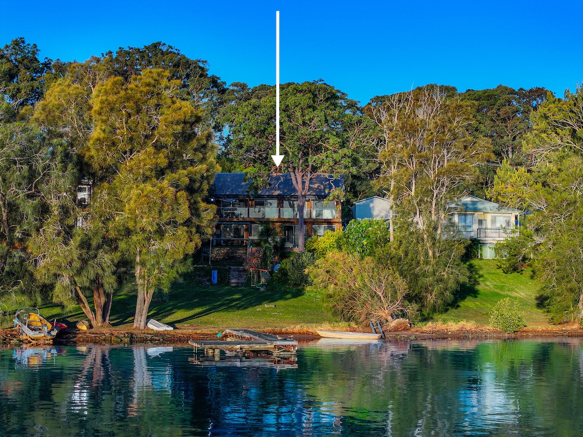 A lakeside view showcases a multi-level retreat nestled among lush greenery. The building features large balconies and is surrounded by trees, adding to the serene environment. Reflections of the structure can be seen in the calm water, enhancing the natural beauty of the landscape.