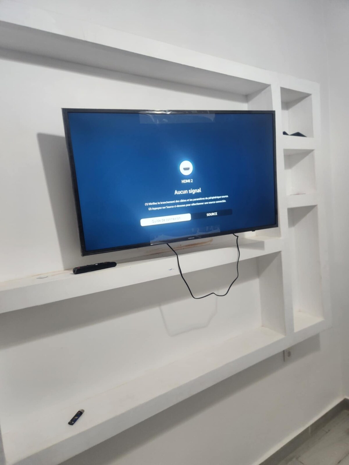 A flat-screen television is mounted on a white feature wall. Below the television, a sleek white shelf is visible, designed for additional storage or display. The overall color scheme is neutral, contributing to a minimalist design.