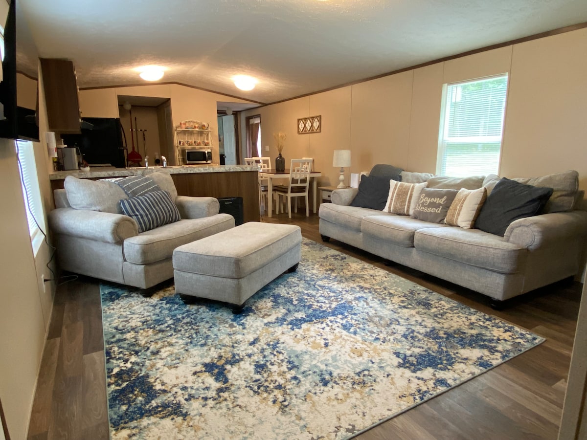 A living area is presented with a light gray couch and an oversized chair, accompanied by a matching ottoman. A plush area rug with a blue and cream pattern covers the floor. Natural light enters through two windows, illuminating the space.