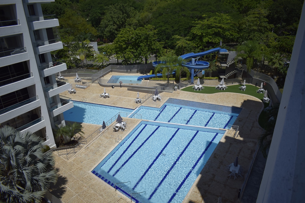 The outdoor pool area features multiple swimming pools, including a large water slide. Surrounding lounge chairs are arranged for sunbathing, and green trees provide shade in the background. The environment suggests a family-friendly atmosphere conducive to relaxation and leisure.