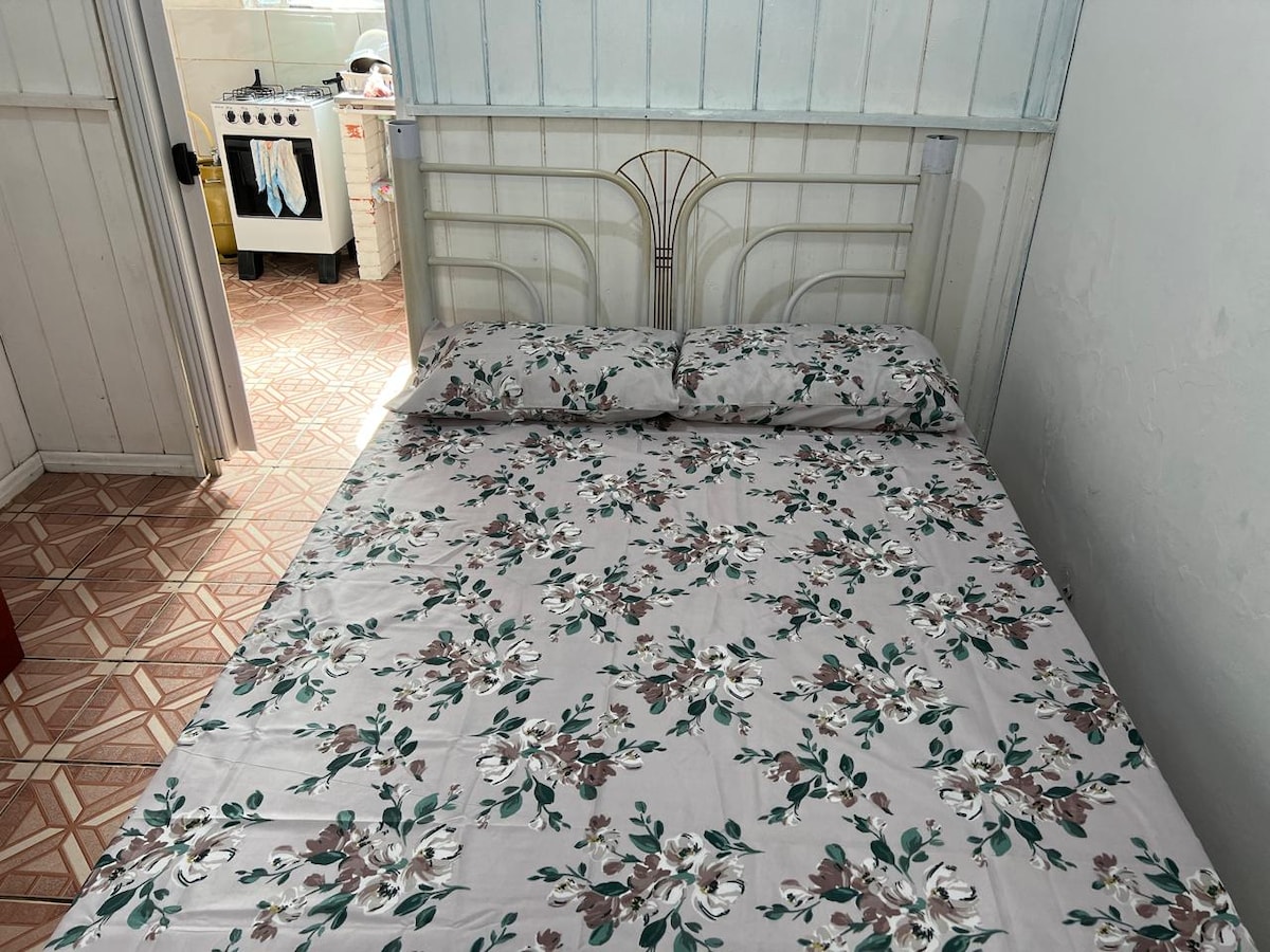A neatly made bed features floral-patterned bedding. The room is illuminated by natural light, creating a bright atmosphere. In the background, the kitchen area is visible, showcasing a stove and cabinetry, contributing to the overall functional layout of the space.