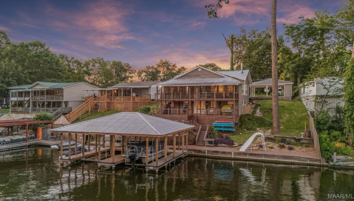 A tranquil lakefront view captures a modern house with a spacious deck. The property features a private dock with boat lifts and lush greenery surrounding the area. Soft colors of the sunset reflect on the calm water, enhancing the natural beauty of the setting.