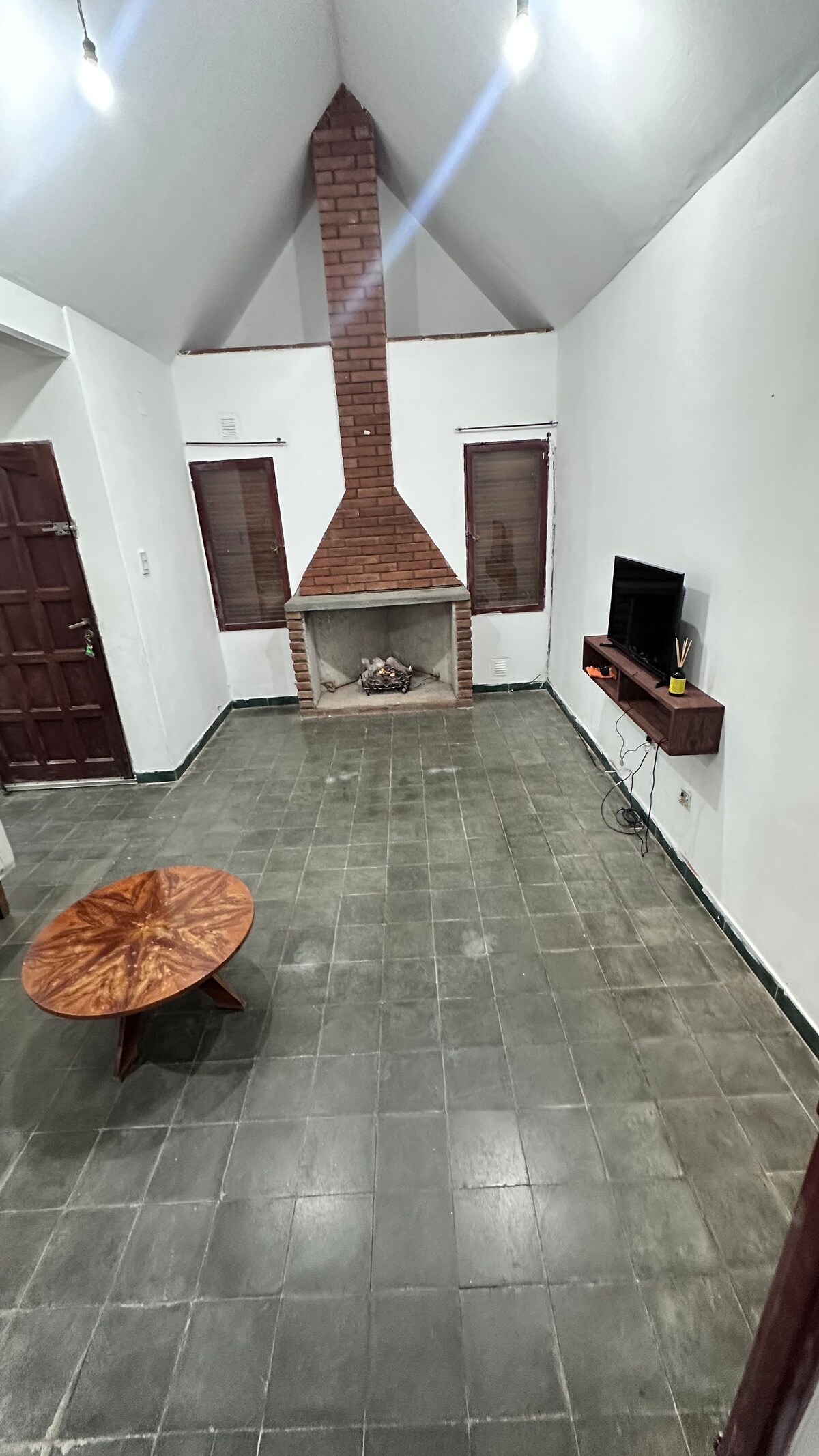 A living room features a rustic brick fireplace centered against a white wall. A wooden coffee table is positioned in front of the fireplace. Flat-screen television is mounted on the wall, with a small shelf for storage beneath. Large windows with wooden shades allow natural light to flow in.