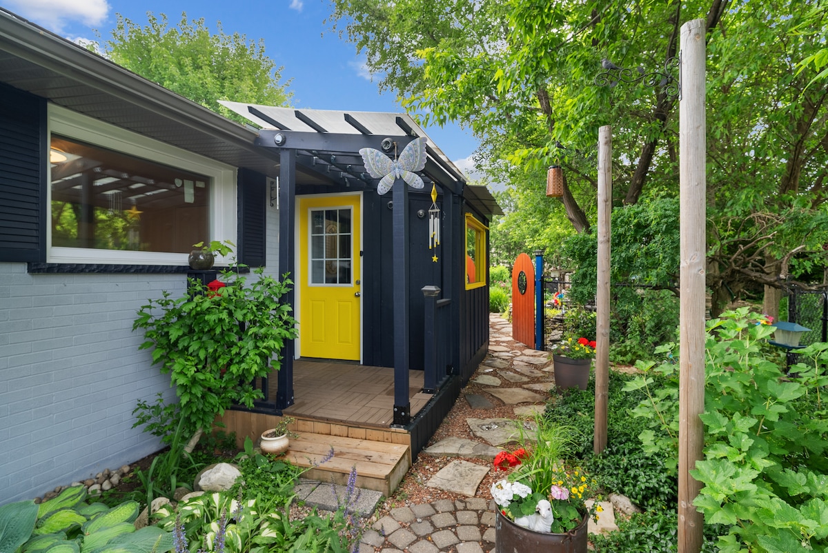 A charming entrance to the cottage features a bright yellow door framed by lush greenery and colorful flowers. A stone pathway leads to the entryway, which is protected by a roof overhang. Decorative elements, such as a butterfly motif and artistic structures, enhance the inviting atmosphere.