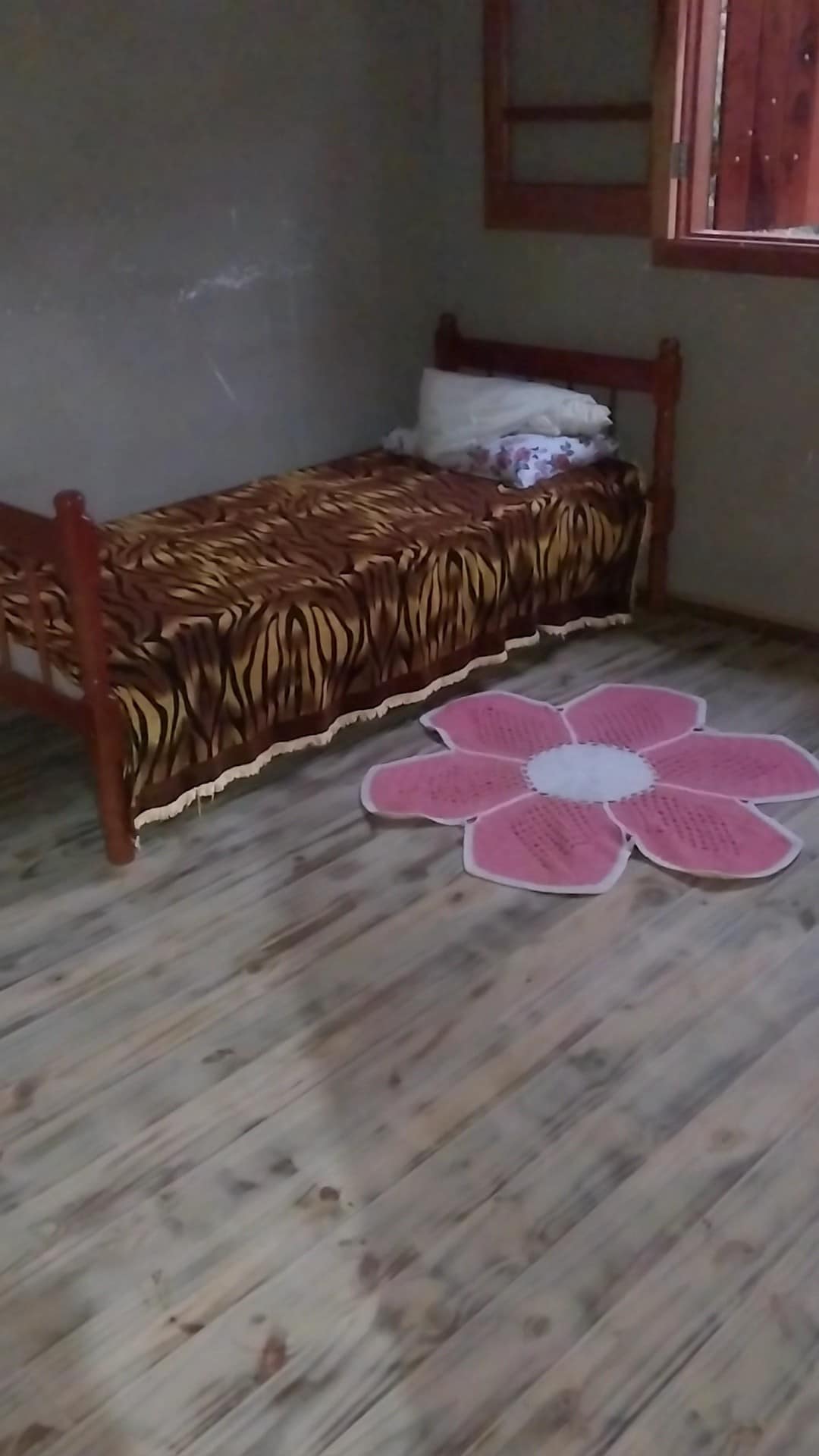 A simple bedroom features a wooden bed adorned with a tiger-striped blanket and a pillow. A colorful flower-shaped rug is positioned on the rustic wooden flooring, adding contrast to the space. Natural light filters in through a window with wooden frames.