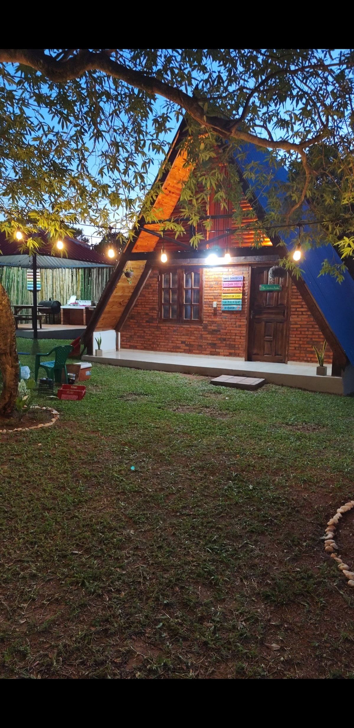 An A-frame structure features warm brick walls and a colorful roof. Subtle lighting enhances the inviting outdoor space, surrounded by well-maintained greenery. A wooden door and framed windows provide a welcoming entrance, while decorative elements emphasize the serene atmosphere of the setting.