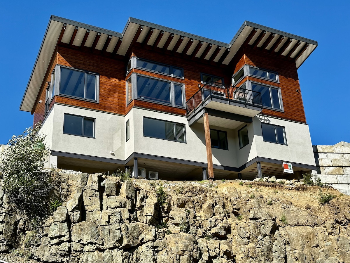 A modern chalet with a combination of wooden and concrete elements is set against a clear blue sky. Large windows allow ample natural light and offer scenic views. The structure is elevated, showcasing a rocky landscape beneath it, enhancing the surrounding natural beauty.