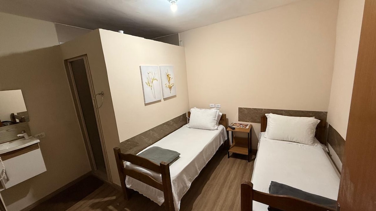 A simple room features two single beds with neutral linens and matching wooden frames. A small bedside table is positioned between them, while a mirror and wall decor enhance the space. Light walls and wood flooring contribute to a calm atmosphere.