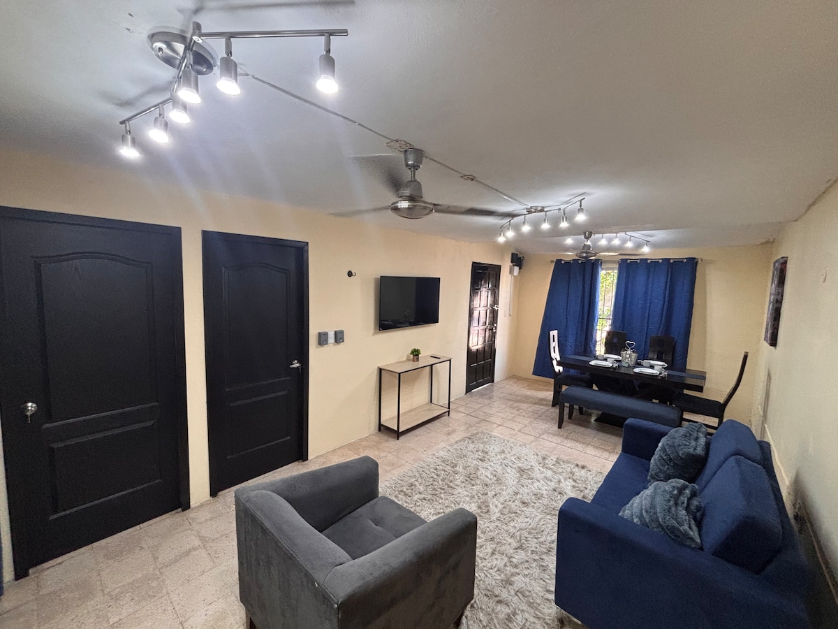 A living area is presented with a gray rug covering the floor. A dark blue sofa and a gray armchair are positioned towards a black coffee table. A wall-mounted TV is visible, alongside two closed black doors, and a dining area with a black table and white chairs is in the background.