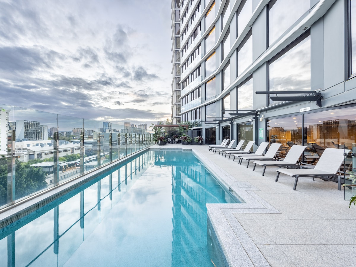 An outdoor swimming pool is situated on a rooftop, offering a view of the surrounding city skyline. Lounge chairs are lined along the pool's edge. The area is surrounded by glass railings, and the atmosphere is enhanced by a soft, cloudy sky.