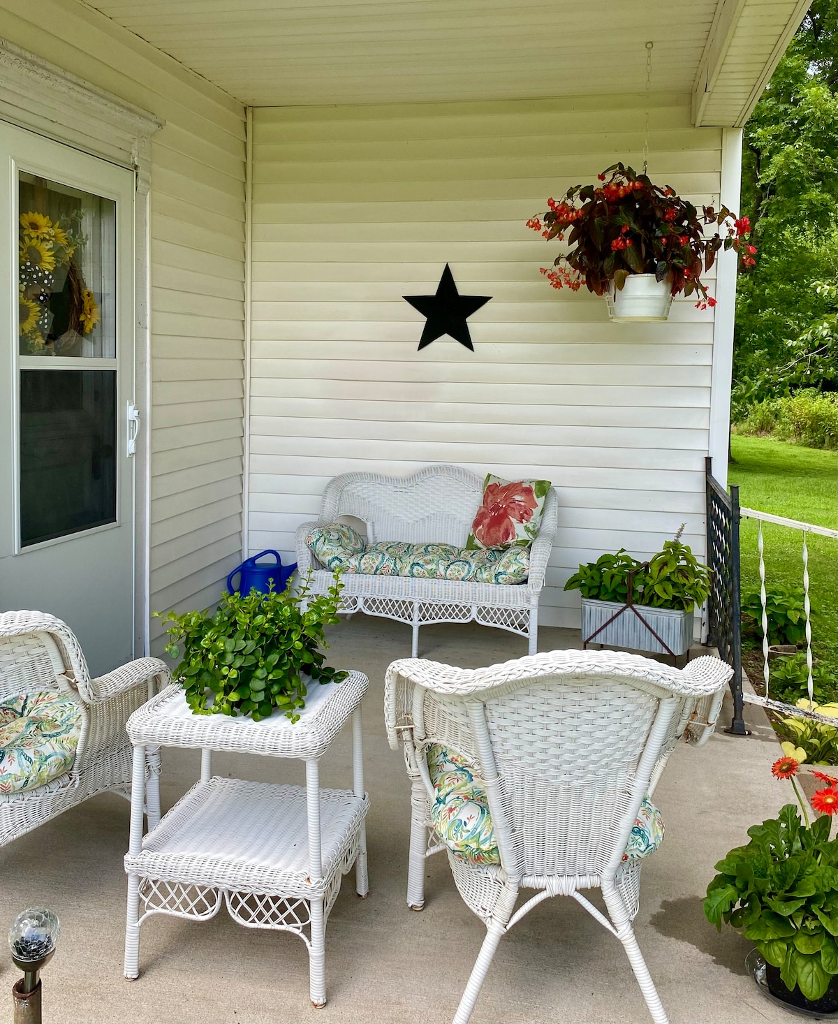 A cozy outdoor seating area is presented with white wicker furniture, including two chairs and a small table, adorned with colorful cushions. Lush green plants are placed around, and a decorative black star is hung on the wall, providing a welcoming touch to the space.
