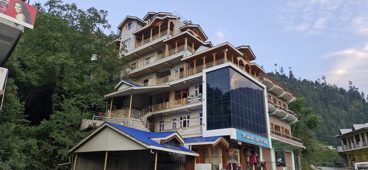 The three-story hotel building features a combination of wooden balconies and glass façades. Lush greenery surrounds the property, complementing its architecture. The entrance is marked by a covered porch, inviting guests into a welcoming environment.