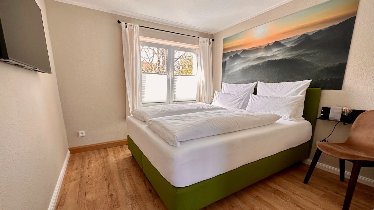 A well-appointed sleeping area features a spacious bed with crisp white linens, flanked by soft pillows. Natural light filters through two side-by-side windows adorned with light curtains. A wall-mounted television and a wooden chair add functional touches to the warm-toned room.