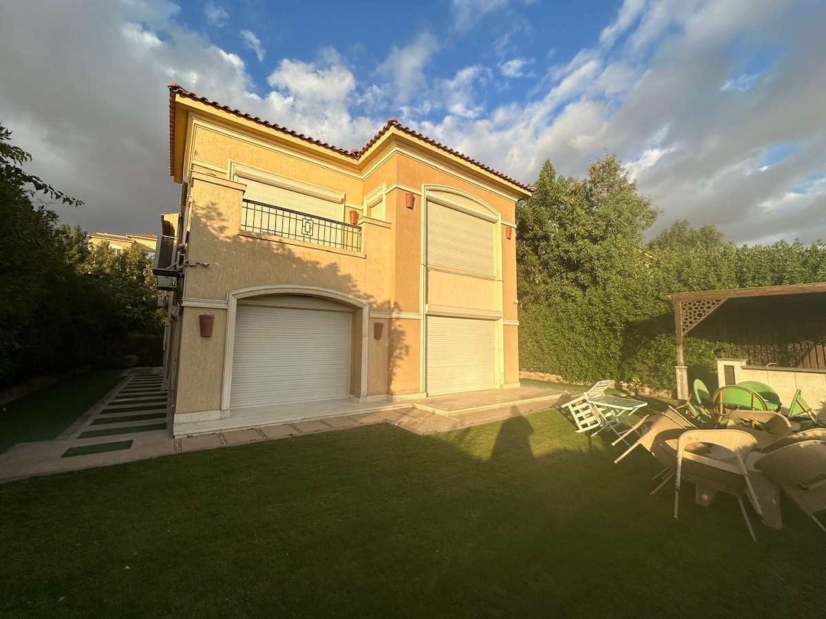 An elegant two-story exterior of a house is presented, showcasing a well-maintained garden with lush greenery. Two large garage doors are visible, alongside a covered outdoor seating area with multiple chairs arranged around a table, providing ample space for relaxation.