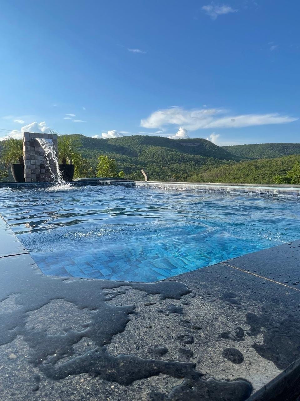 A glistening swimming pool is framed by natural stone features, with a gentle waterfall cascading into the clear water. Lush green hills rise in the background under a bright blue sky, creating a serene and inviting outdoor space.