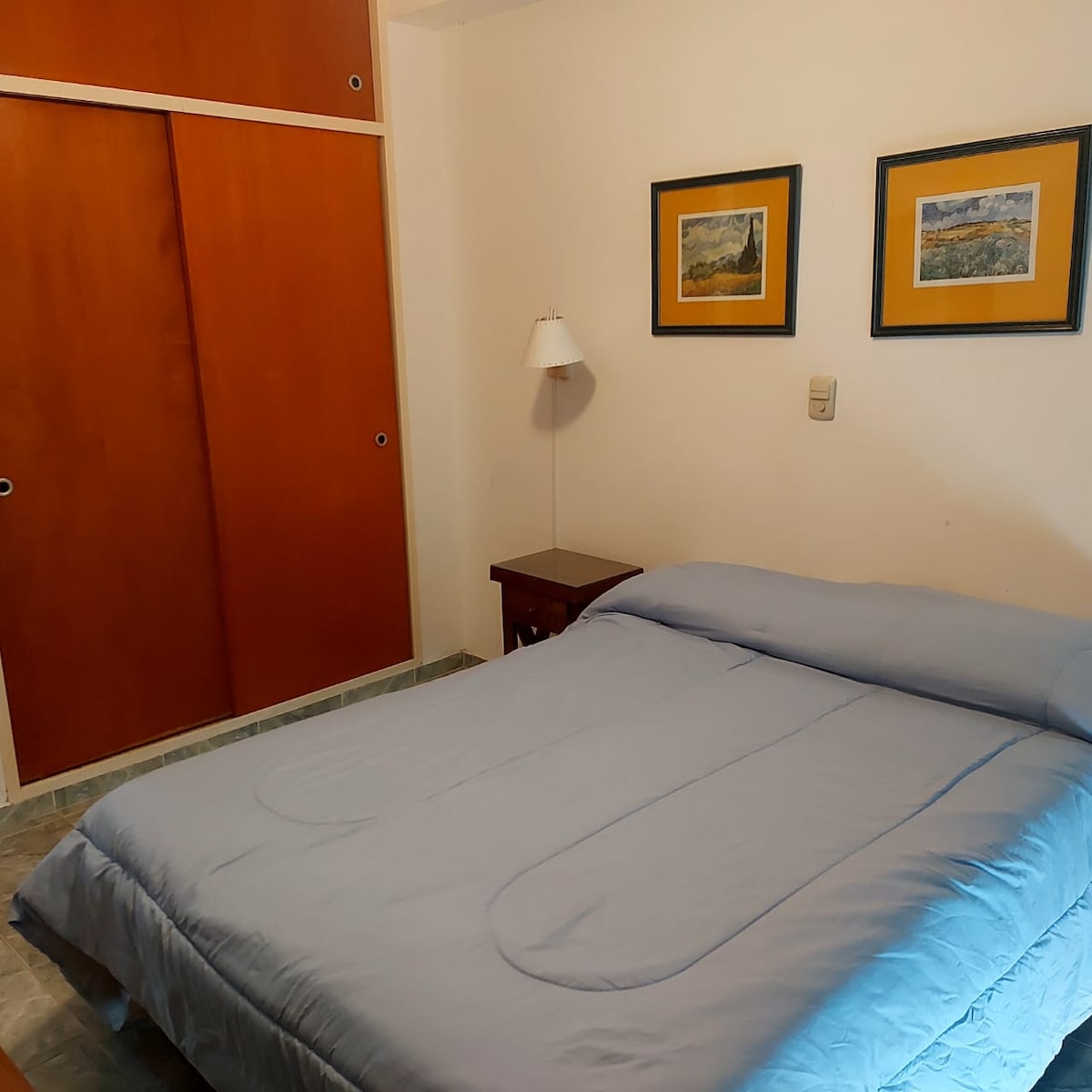 A cozy sleeping area features a spacious bed with a light blue blanket. Framed artwork adorns the walls, providing subtle color. A bedside table and a lamp are positioned beside the bed, while a wooden closet is visible in the background.
