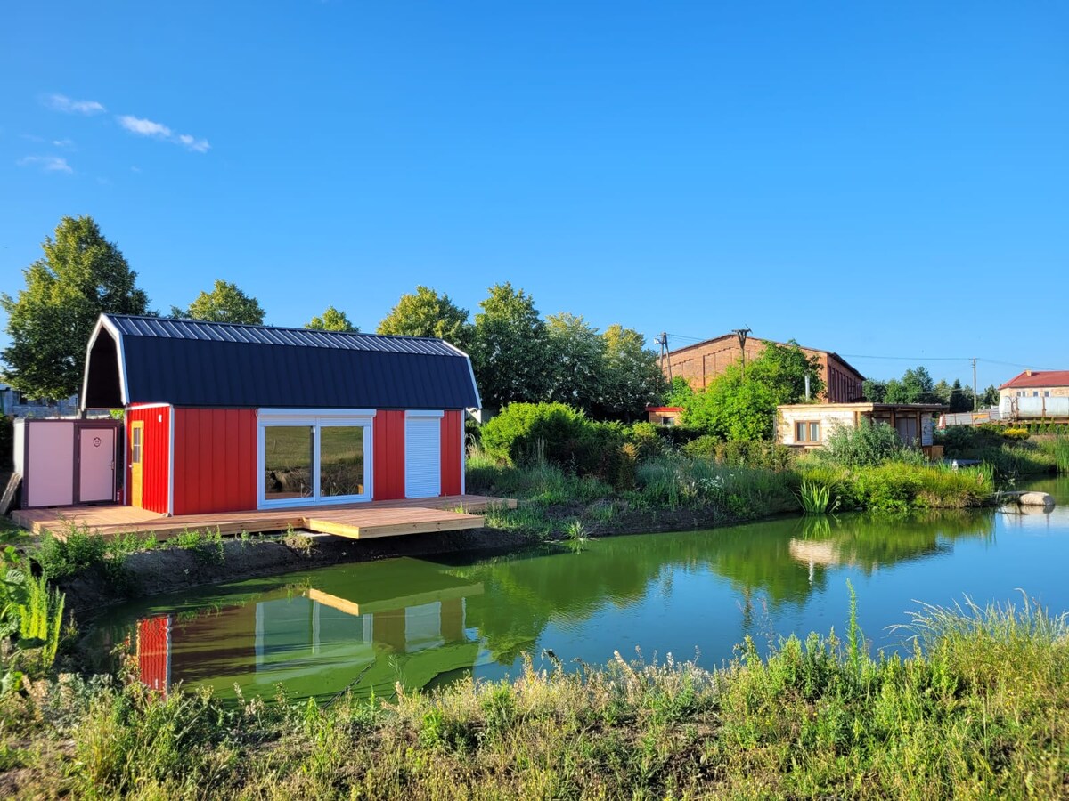 A charming red and white house is situated adjacent to a serene pond. Lush greenery surrounds the area, with reflective waters visible in the foreground. The structure features a sloped roof and large glass windows, providing views of the vibrant outdoor landscape.
