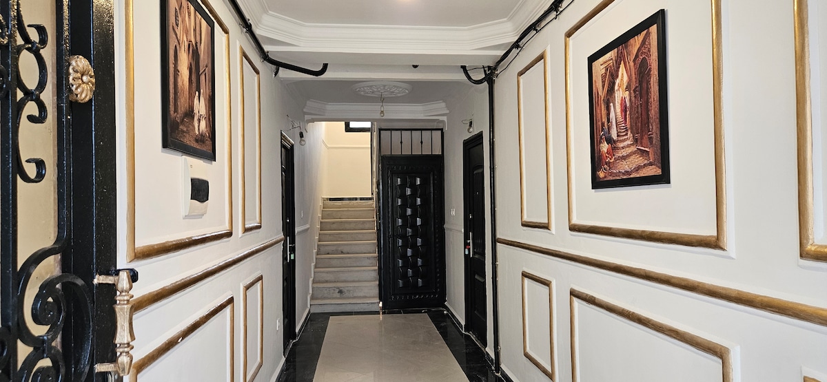 The hallway features elegant walls adorned with framed artwork, complemented by gold trim. A polished black and white marble floor leads toward a staircase that is partly visible, while dark doors are positioned on either side, providing access to individual rooms.
