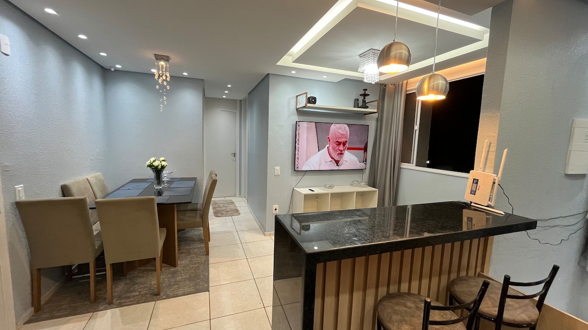 A contemporary living space features a dining table set for six, complemented by beige chairs. A kitchen island with a dark countertop separates the dining area from the living space, where a television is mounted on the wall. Soft lighting enhances the neutral color scheme.